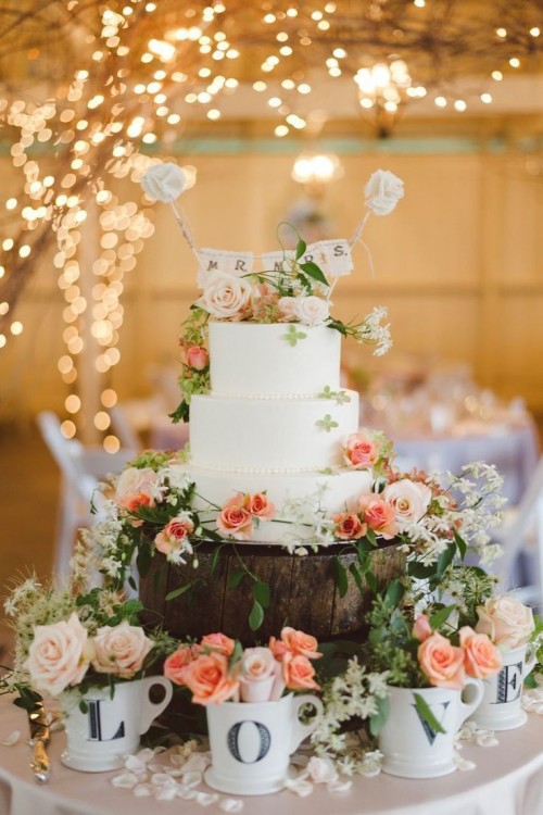 a white spring wedding cake decorated with blush, peachy blooms and greenery, a banner and white fabric blooms