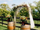a chic vineyard wedding arch of metal, with neutral fabric, with neutral and bright blooms and greenery is a chic idea