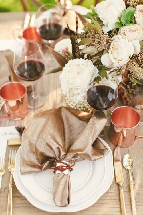 an elegant neutral fall wedding tablescape with shiny linens, copper mugs, a neutral floral centerpiece and elegant gold cutlery