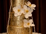 a shiny gold patterned wedding cake topped with white sugar blooms and pearl strands is a chic idea with a refined touch