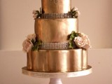 a shiny gold wedding cake with embellishments, blush blooms and leaves looks super refined and chic