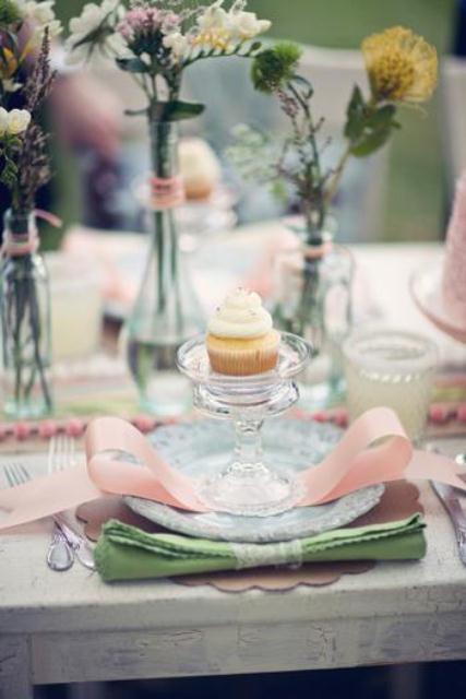 a pastel spring wedding table with wildflowers in bottles and vases, green napkins and pink bows plus cupcakes for each place setting