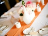 a bright spring wedding tablescape with an orange runner and ribbons, bold blooms in white vases and candles plus neutral porcelain