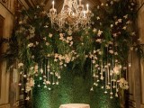 a greenery backdrop with greenery and white blooms hanging down and a chandelier for a sophisticated wedding