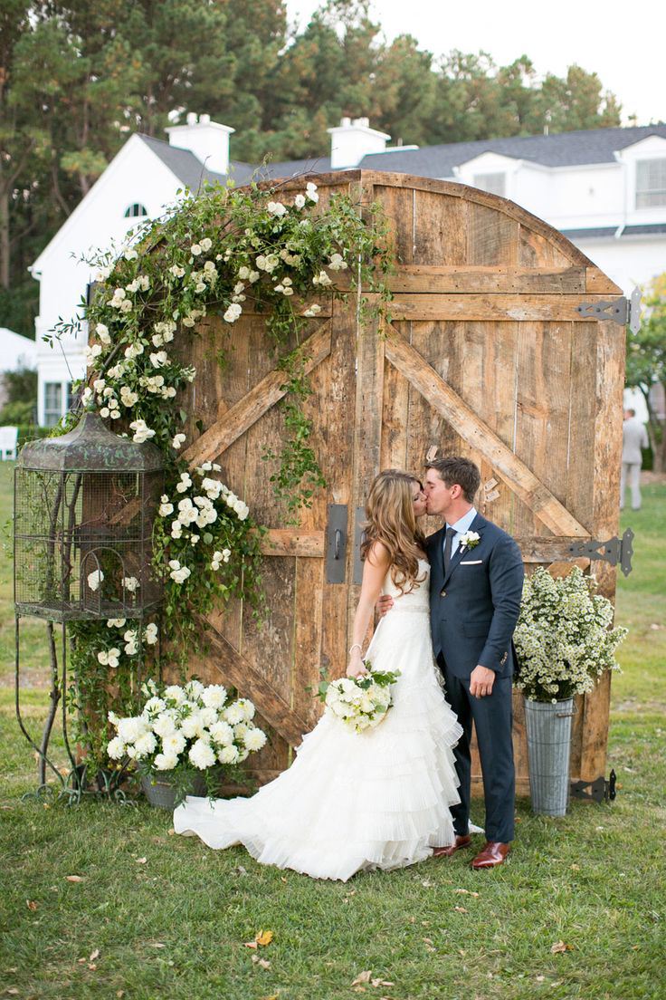 Old barn doors with greenery and white blooms, a cage and a bucket with blooms is a chic wedding backdrop
