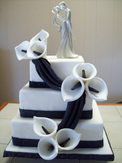 a white square wedding cake decorated with black ribbons, sugar callas and a topper