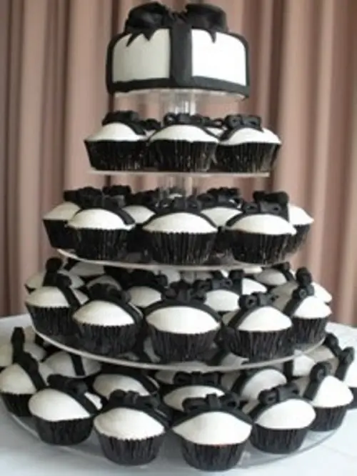 a simple white wedding cake decorated with a black sugar ribbon and bow and lots of matching cupcakes