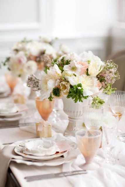 a creamy and pastel spring wedding tablescape with white porcelain, neutral napkins, creamy and blush centerpieces with greenery, peachy glasses