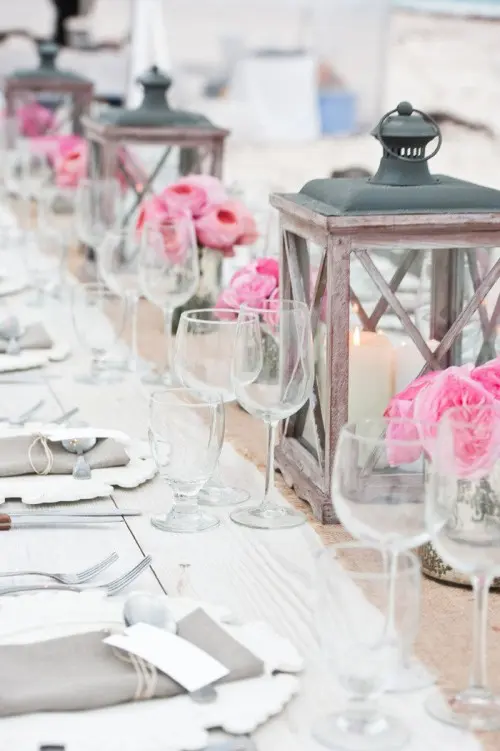 a vintage wedding tablescape with a burlap table runner, pink peony roses, wooden candle lanterns, grey napkin and porcelain plates
