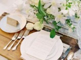 a neutral spring wedding tablescape with white blooms and greenery, a white table runner and white plates, silver cutlery