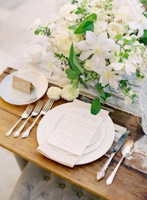 a neutral spring wedding tablescape with white blooms and greenery, a white table runner and white plates, silver cutlery
