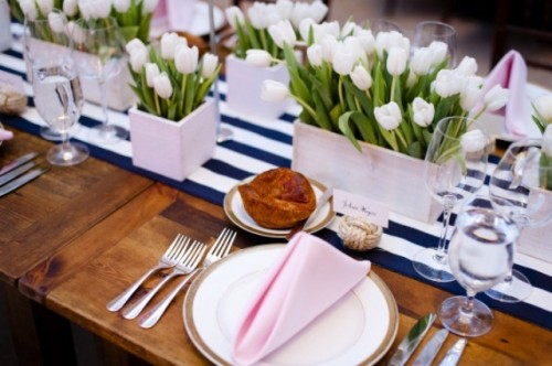 a spring wedding tablescape with a navy and white table runner, white tulips, white porcelain and pink napkins plus silver cutlery