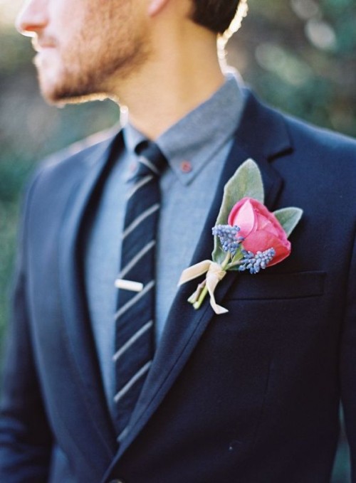 a navy woolen suit, a grey shirt, a striped navy and white tie and a bright floral boutonniere