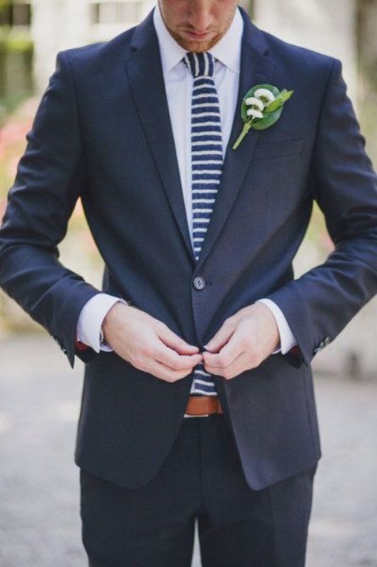 A navy suit, a white shirt are spruced up with a navy and white striped tie and a catchy boutonniere