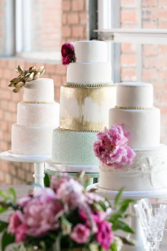white cake stands and an assortment of various pastel and metallic wedding cakes with pink blooms