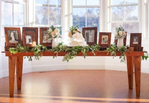 a sweets wedding table with greenery and family photos of people who are gone but loved and white wedding cakes