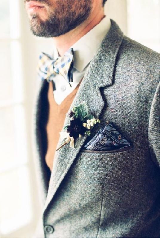 A chic boho groom's outfit with a white shirt, bold printed bow tie, a tan waistcoat, a grey woolen blazer, a dark printed handkerchief and a floral boutonniere is a stylish idea