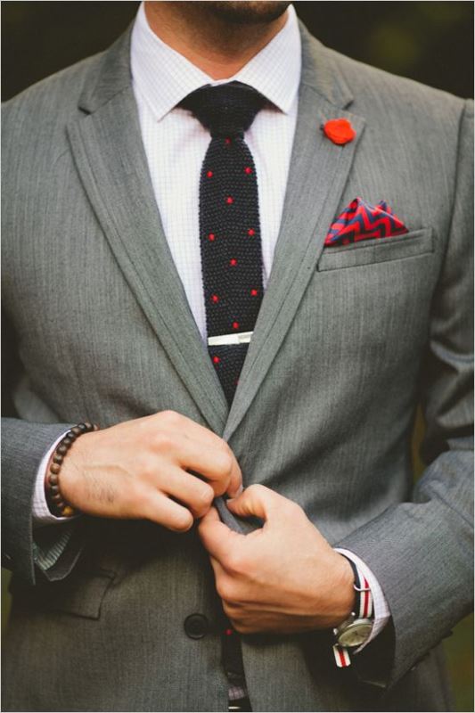 A bold and playful groom's outfit with a grey suit, a white shirt, a black polka dot tie, a bold red printed handkerchief and a red badge as a boutonniere