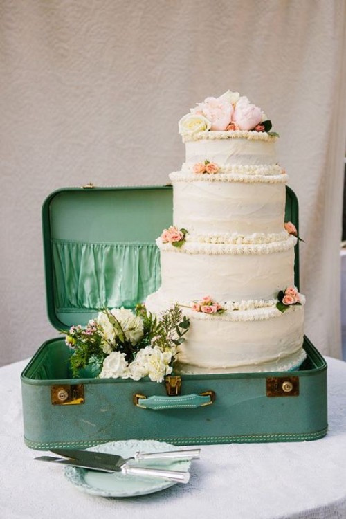 a green vintage suitcase repurposed into a cake storage unit, with a white cake with some pink blooms and greenery and a white floral arrangement is a lovely idea