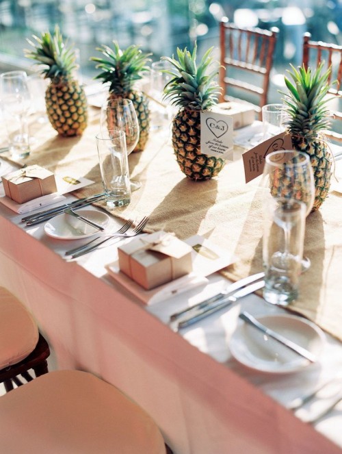 a neutral tropical wedding tablescape with a burlap runner, white porcelain and silver cutlery, pineapples are given as wedding favors