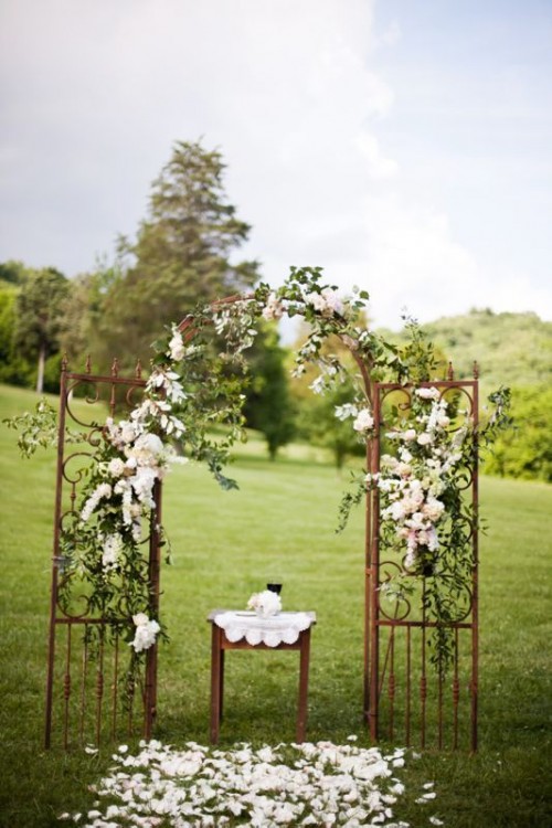 a rusty metal wedding arch decorated with lush greenery and white blooms looks sophisticated and beautiful