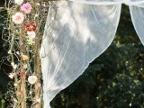a delicate spring wedding arch interwoven with bright blooms and some vines and decorated with white fabric