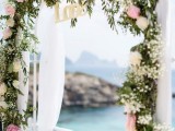 a spring wedding arch decorated with greenery, pink and white blooms, white fabric and the word LOVE