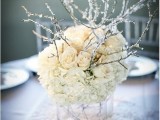 a frozen winter wedding centerpiece of a clear vase, white roses and hydrangeas and branches that seem frozen