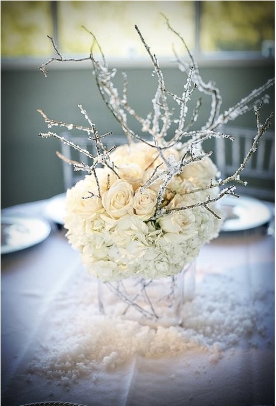 A frozen winter wedding centerpiece of a clear vase, white roses and hydrangeas and branches that seem frozen
