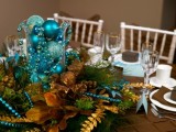 a colorful winter wedding centerpiece of a gilded pinecone, faux evergreens and gold ribbons and turquoise ornaments and beads