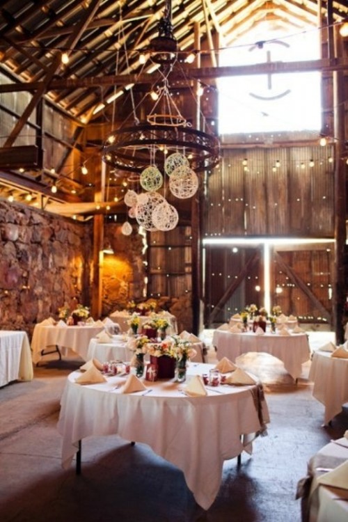 a chic barn wedding reception space with a chandelier with yarn balls, bold blooms, lights and neutral linens is all chic