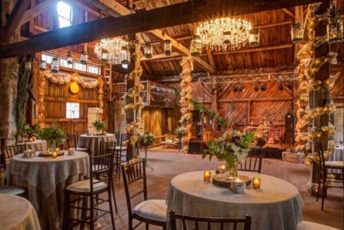 a rustic barn wedding reception space with chandeliers, white blooms, greenery and grey linens is a very welcoming and stylish space