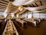 a barn wedding reception space decorated with white fabric curtains, greenery and lights and with tables lined up with burlap