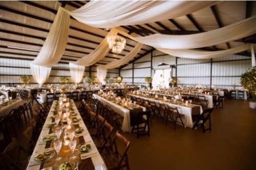 a barn wedding reception space decorated with white fabric curtains, greenery and lights and with tables lined up with burlap