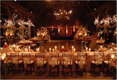 a refined decorated barn wedding reception space with lots of lights, candles, blooms, trees and white fabric banners and garlands