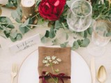 a boho wedding tablescape with greenery, a red bloom, some feathers and candles, an elegant gold-rimmed plate and cutlery plus a neutral napkin with a velvet ribbon bow