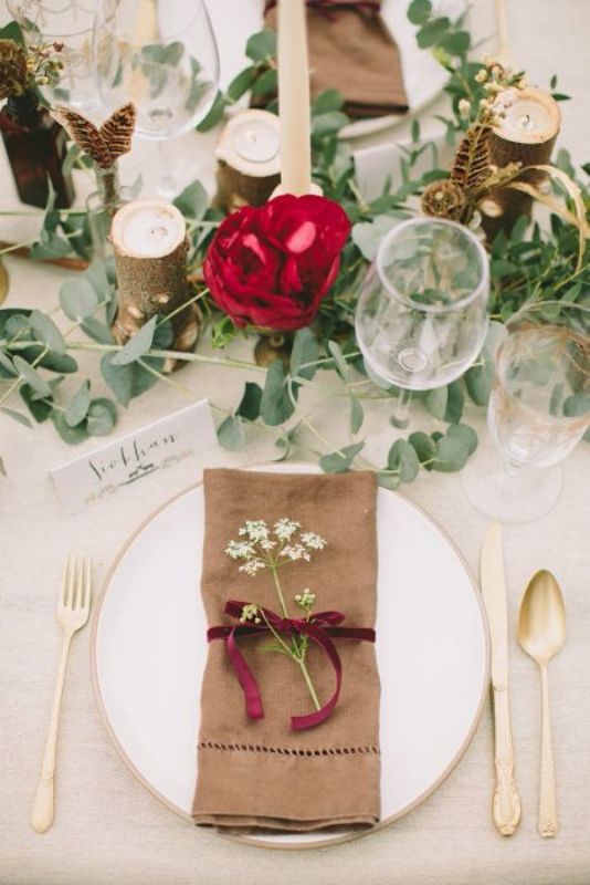 A boho wedding tablescape with greenery, a red bloom, some feathers and candles, an elegant gold rimmed plate and cutlery plus a neutral napkin with a velvet ribbon bow