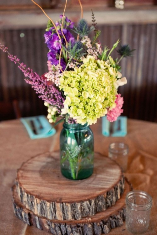 a bright barn wedding centerpiece of a jar with bright purple and yellow blooms placed on wood slices