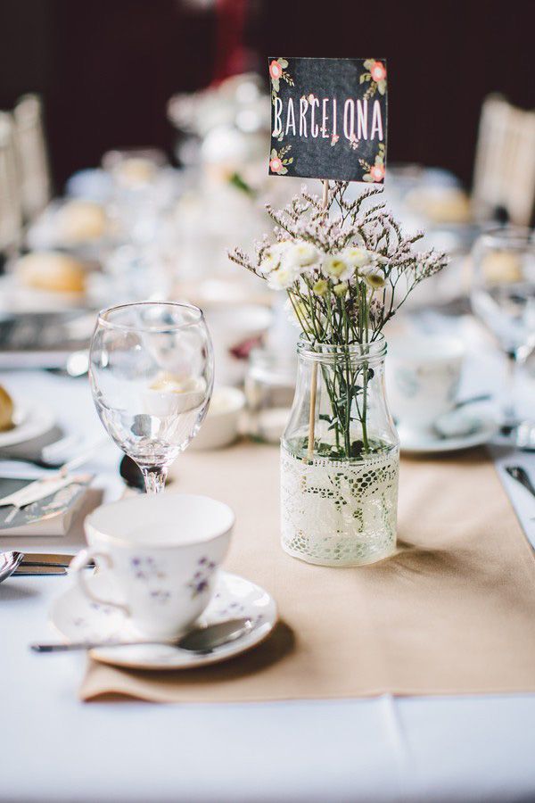 A clear vase wrapped with white lace, with dried herbs and flowers plus a table name