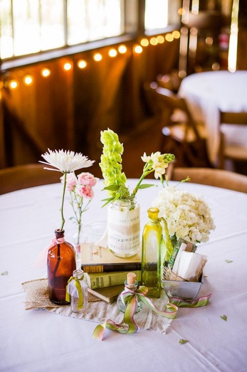 a barn wedding centerpiece with bottles and jars, white, pink and green blooms, a stack of books and some fabric