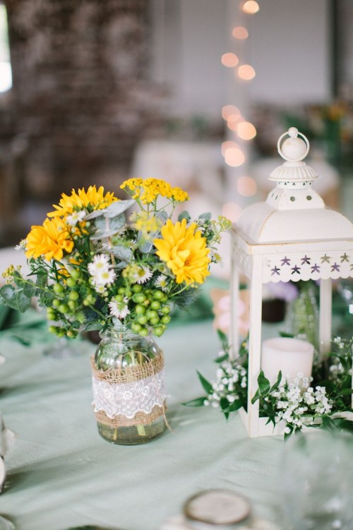 a jar wrapped with burlap and lace and white and yellow blooms, a white candle lantern with greenery and white flowers