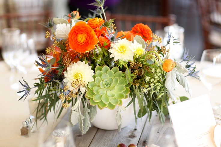 a barn wedding centerpiece of neutral, orange and green blooms, greenery and thistles is super lush