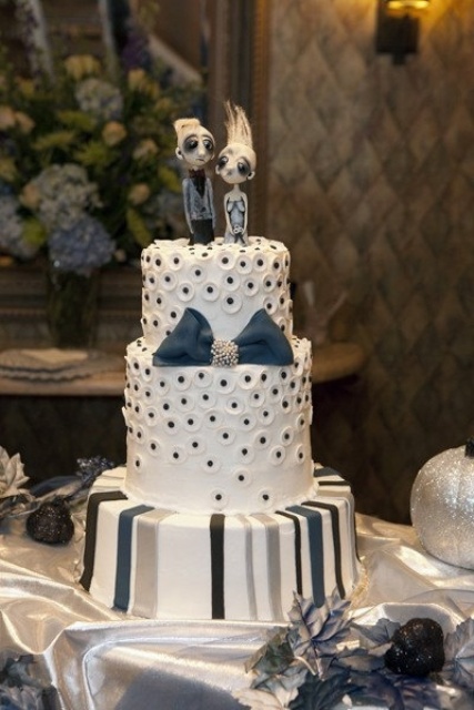 a white, grey and blue wedding cake with various patterns and a blue bow plus quirky toppers for a Halloween wedding
