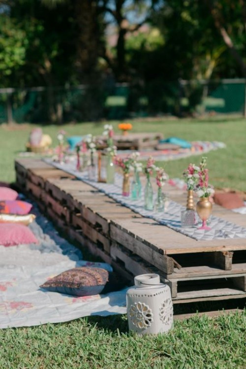 a bright boho picnic setting with a low pallet table, bright blooms, blankets and pillows for a rehearsal dinner