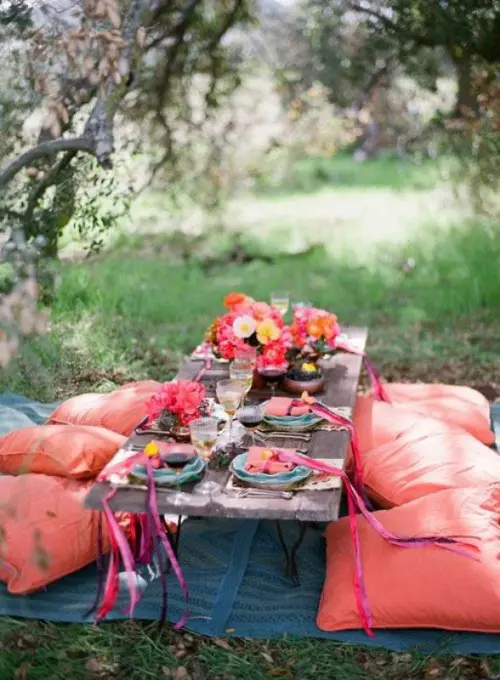 a bright boho themed rehearsal dinner picnic with bold red pillows, bold blooms, printed blankets and lots of delicious food