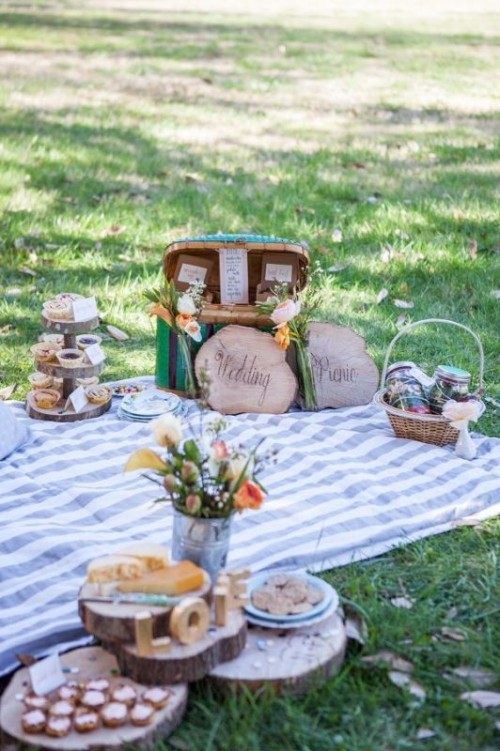 a cozy picnic setting with a striped blanket, some blooms, wood slices, a vintage suitcase and baskets with food and drinks