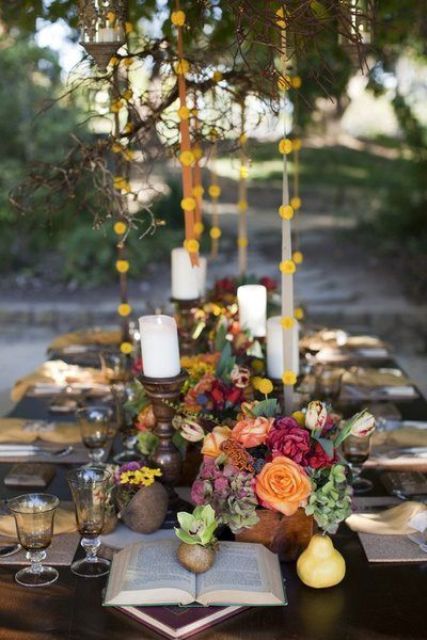 a bright fall wedding tablescape with bold blooms, greenery, fruit, candles in refined candleholders, vintage glasses and books