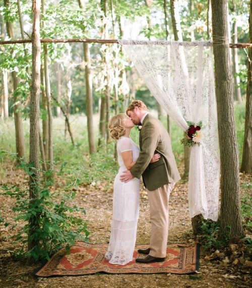 a woodland wedding ceremony space with a wedding arch made on a living tree, with a lace curtain and a boho rug plus bold blooms