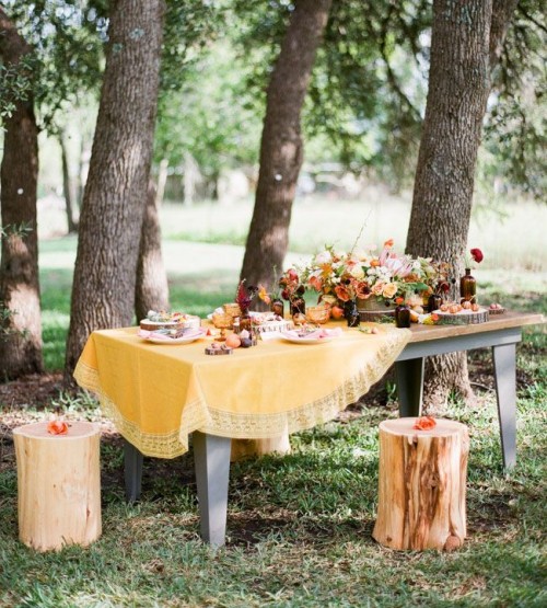 a bright fall woodland wedding reception with a mellow yellow tablecloth, bold blooms and dark glass vases and jars, greenery and tree stumps as seats