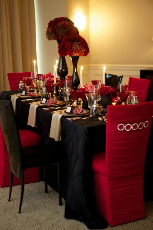 a red and black wedding tablescape with a black tablecloth, black and red chairs, black vases with red roses